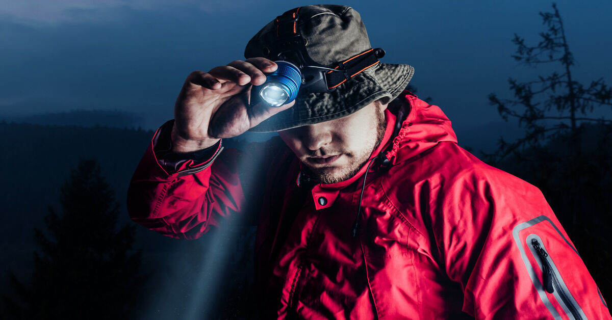 A man in a red jacket looking down as he adjusts the headlamp attached to his bucket hat.